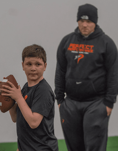 Young boy practicing football with coach watching.